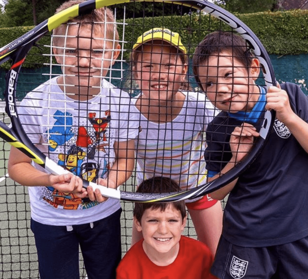 4 young tennis players standing together on an AstroTurf court behind a huge tennis racket after playing mini tennis at Bath Tennis